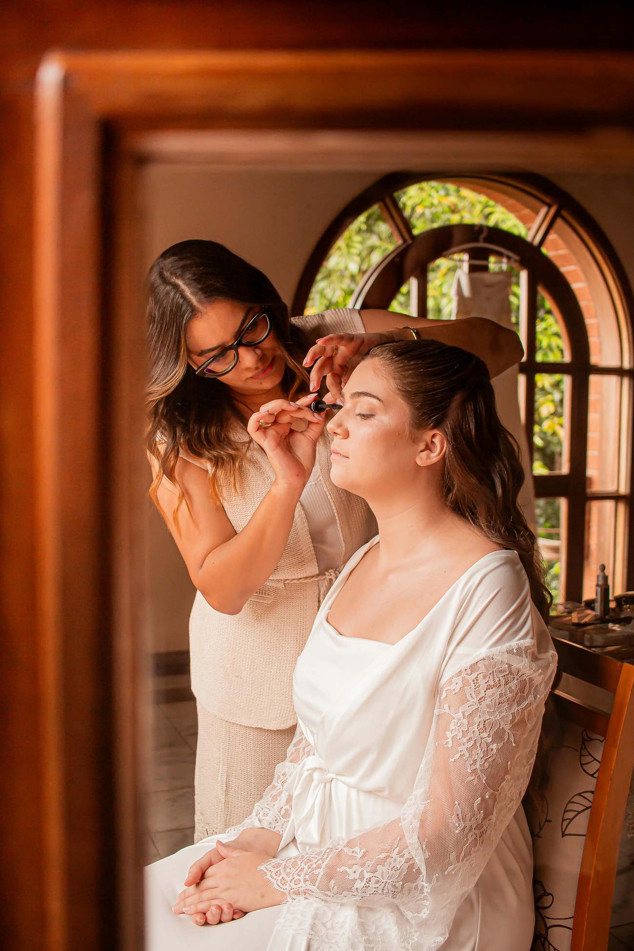 Maquiadora aplicando rímel em uma cliente de robe de renda diante de uma janela em arco
